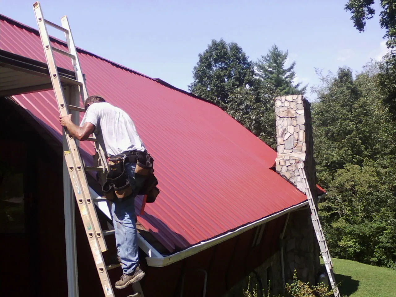 Roofer on a ladder beside a red metal roof and stone chimney — Soffit & Fascia Repair by Roofing Brothers in Flinton